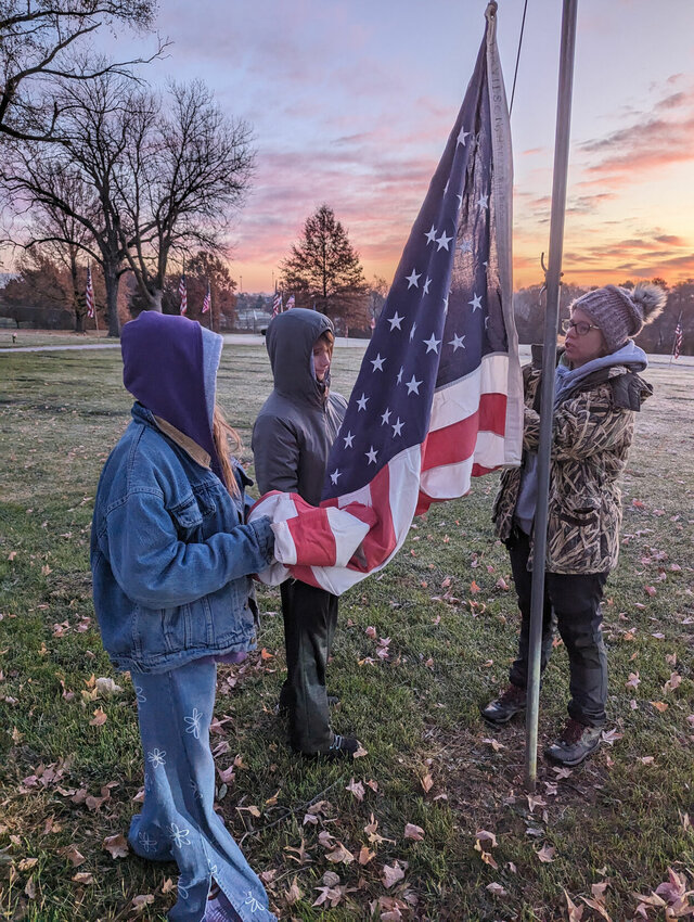 Elizabeth Foerster, Gillian Grogan, and Scoutmaster Christine Steele hoist flags at the cemetery at sunrise on Veteran&rsquo;s Day.