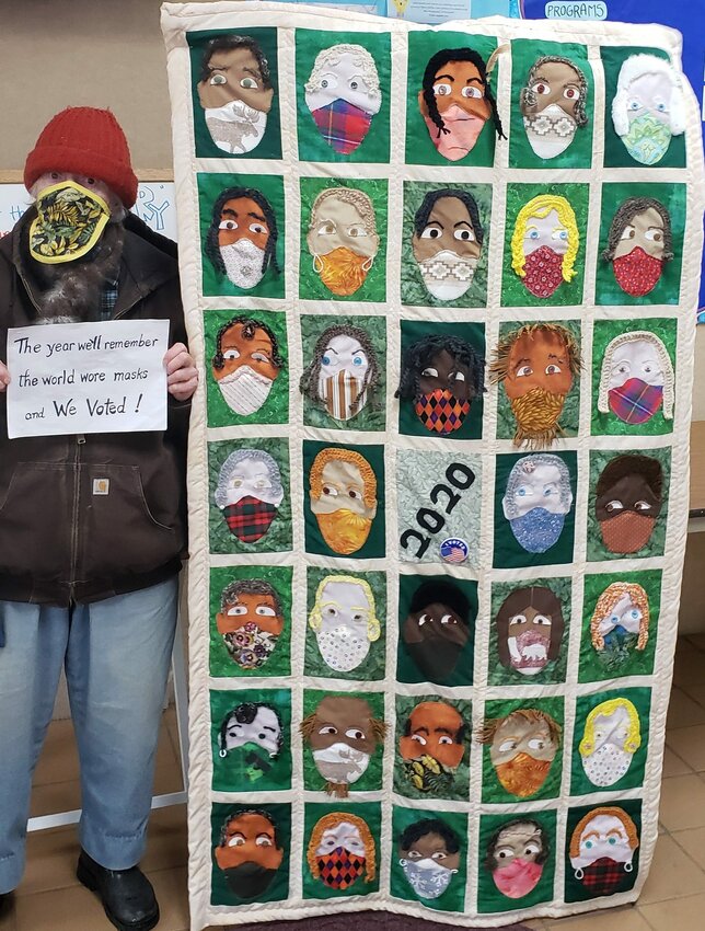 A masked Margaret Naysmith stands with the quilt she made depicting faces of different races all wearing masks which is on display at the Algoma Public Library. Naysmith is holding a sign that normally is next to the quilt explaining her idea behind making it.