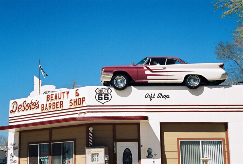 A full-size, brightly painted old DeSoto automobile rests on top of the front of the DeSoto Barbershop building against a backdrop of clear blue sky in Ash Fork, Arizona (Leslie Connell/National Archives).