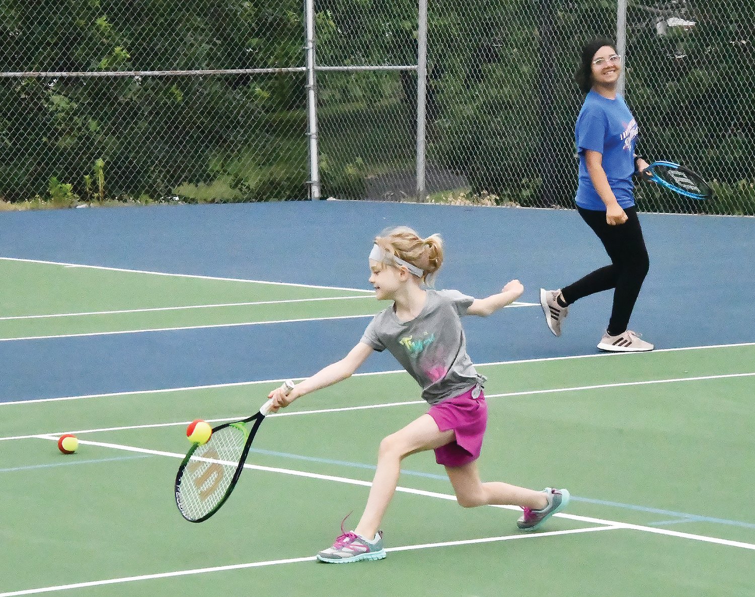 Alexa Moore gets the racket on the ball while Natalie Perez smiles in the background during tennis lessons.