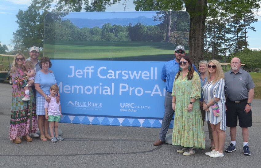 Members of the late Jeff Carswell's family stand with the banner revealing the new name of UNC Health Blue Ridge's annual golf tournament. Pictured are Carswell's wife, Janet Carswell; his sister Sandy Lonon with her husband Rodger and their daughter Kristen Gaston; his daughter Kaitlin Bensch with her husband Jonathan and their children Wells and Grace; and his daughter Keri Jo Carswell with her fiancé Connor Clark.