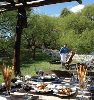 Young lamb is stretched across a traditional iron rack and grilled over an open air fire at a picnic site. Accompaniments are roasted vegetables, breadsticks and the wines of Argentina.