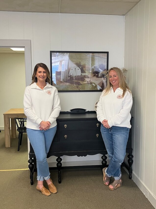 Realtors Courtney Luzinski (left) and Alicia Soper next to a picture of the white barn which serves as inspiration for their business name.