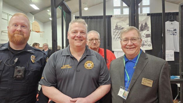 O-W Police Chief Patrick Fehlman, Sheriff Kerry Kirn, and Allan Hodnett in front of the Owen Centennial Committee&rsquo;s Jail &amp; Bail booth. Owen Mayor Scott Jalling is behind bars.