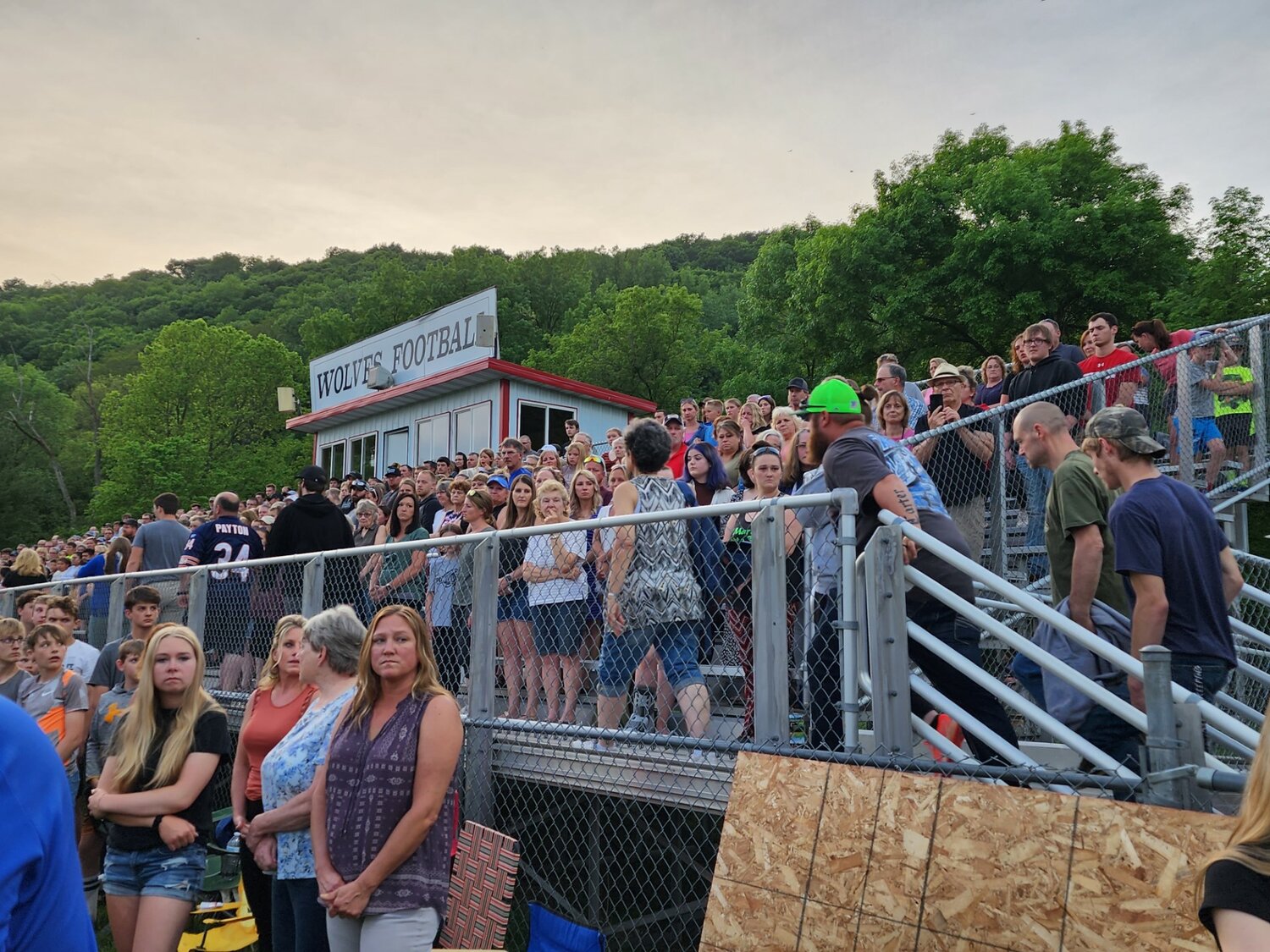 Prayer vigil attendees stand silently as members of the Bleskacek and Loga families file onto the bleachers at a May 31 prayer vigil in Elmwood.