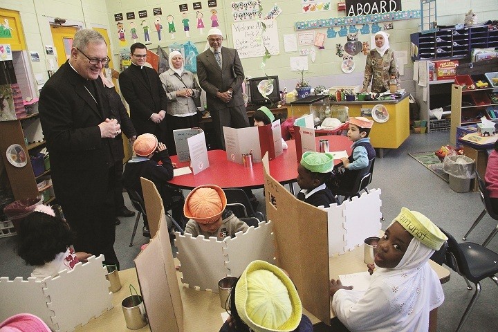 UNITED THROUGH FAITH:   Bishop Thomas J. Tobin reviews some of the work done by Sister Paula (at far right) and her pre-k students at the Islamic School of Rhode Island during his visit to the West Warwick school last week. The students are wearing sailor hats to represent a nautical journey they are making around the United States as they learn about the nation&rsquo;s different regions. Principal Basima Shiladeh Al-Jallad said the school currently has 121 students from across the state enrolled in grades pre-k through eight, and that many graduates go on to pursue their secondary education at one of the Catholic high schools in the diocese. The Islamic School was established in 2004 in a building that housed the former Sacred Heart Catholic school. The Bishop was invited to visit the school by Imam Farid Ansari, president of the Rhode Island Council for Muslim Advancement, pictured at center above with his wife Naima, following a recent meeting he had with a group of Muslim leaders in which they pledged their support for traditional marriage. During the Year of Faith, the U.S. bishops are advancing a movement for life, marriage and religious liberty and seek to share that message with others of faith across the nation.